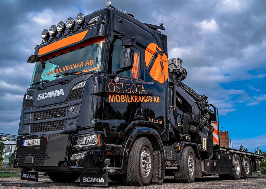 En svart lastbil med orange företagstryck, blå himmel i bakgrunden.