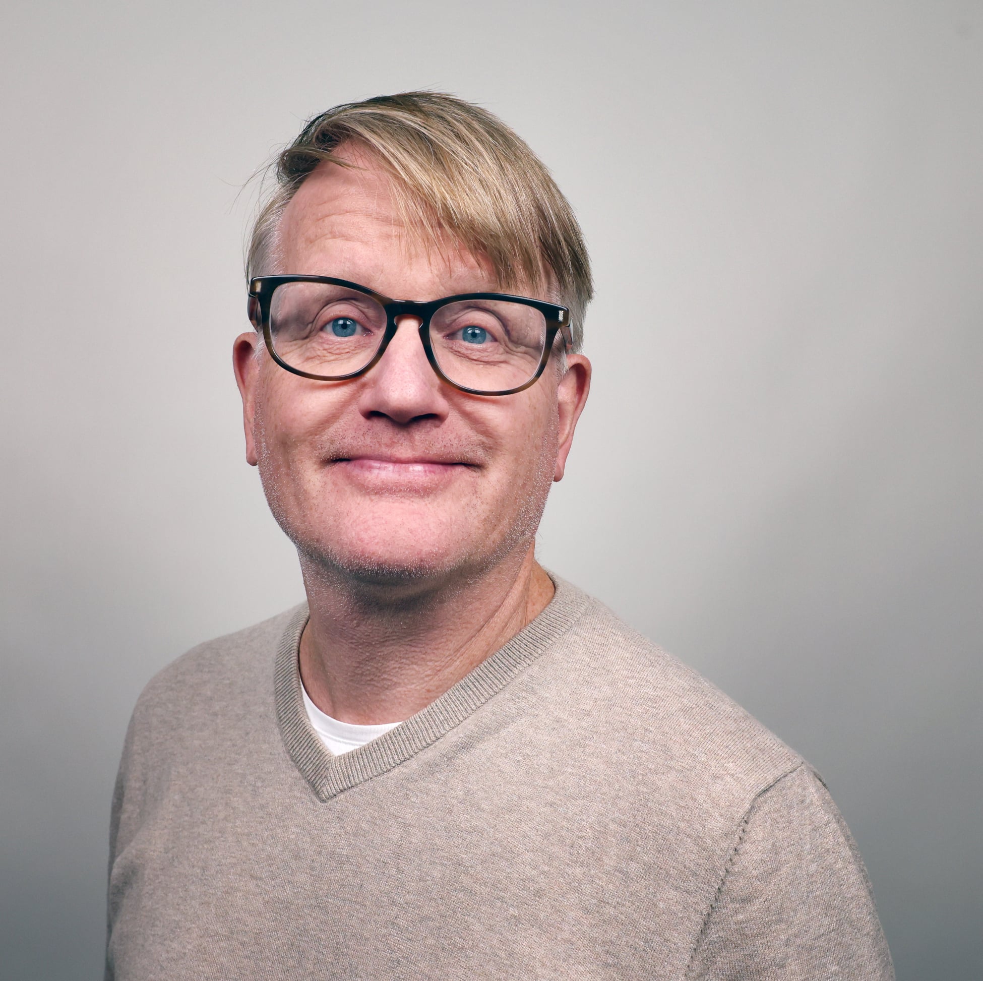 Porträtt på Per Faxér, en man med blont kort hår, glasögon och en beige tröja över en vit t-shirt.